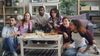A group of people gathered around in a living room with pizza and beers. Two of them are sitting on a couch, holding controllers and focused. The others are watching the game