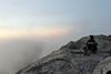 Em Halberstadt, a white woman with dark hair pulled into a half bun, sits cross-legged on a rocky mountaintop, looking out over a foggy expanse.