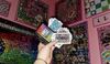 A hand holding up several bedazzled cards from board games like Sorry and Hues and Cues. The wall behind the person's hand is covered in bedazzled board games that have been hung up as art.
