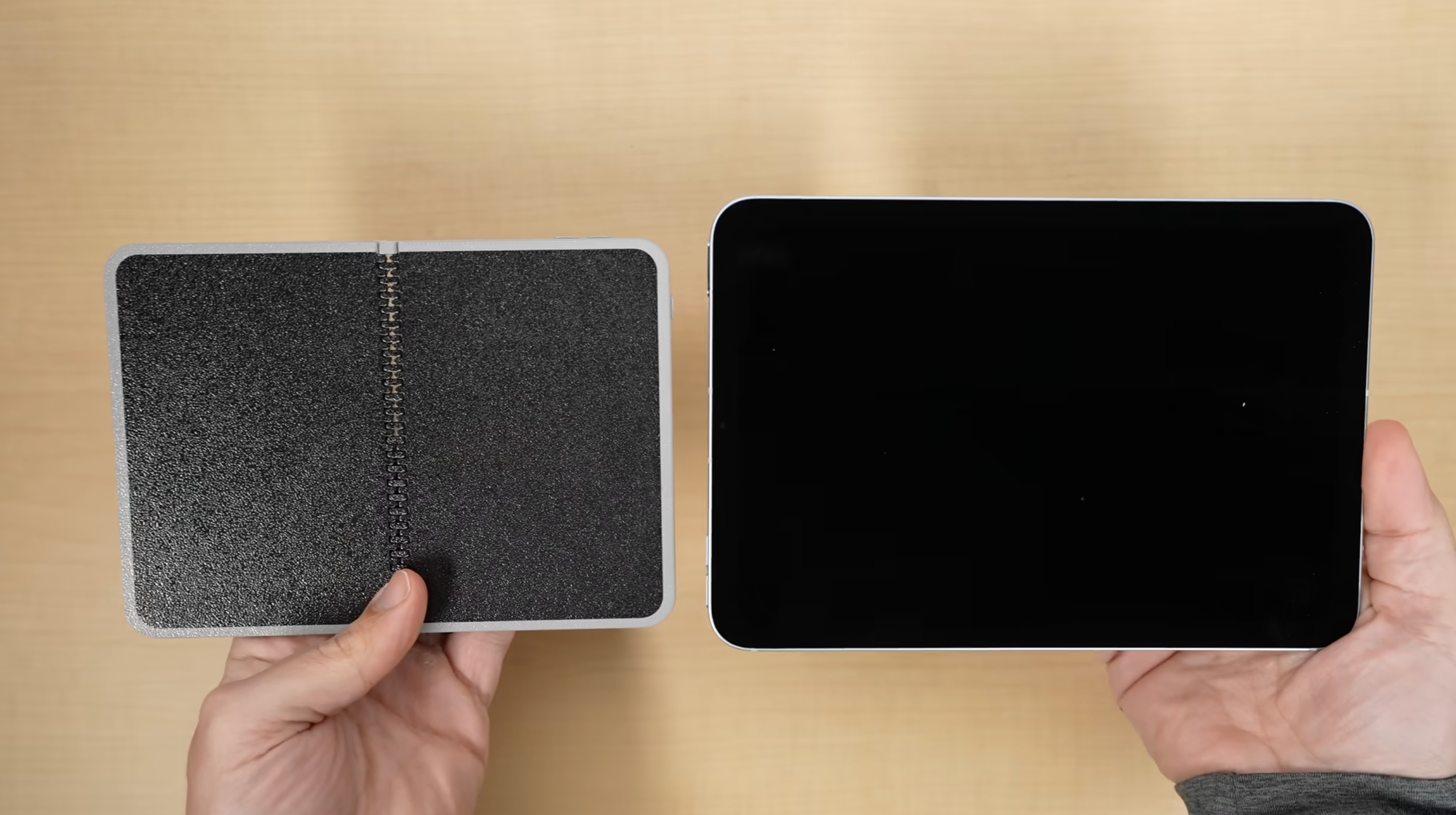 A pair of hands holds the plastic mockup, now open, and a real iPad mini above the same table.
