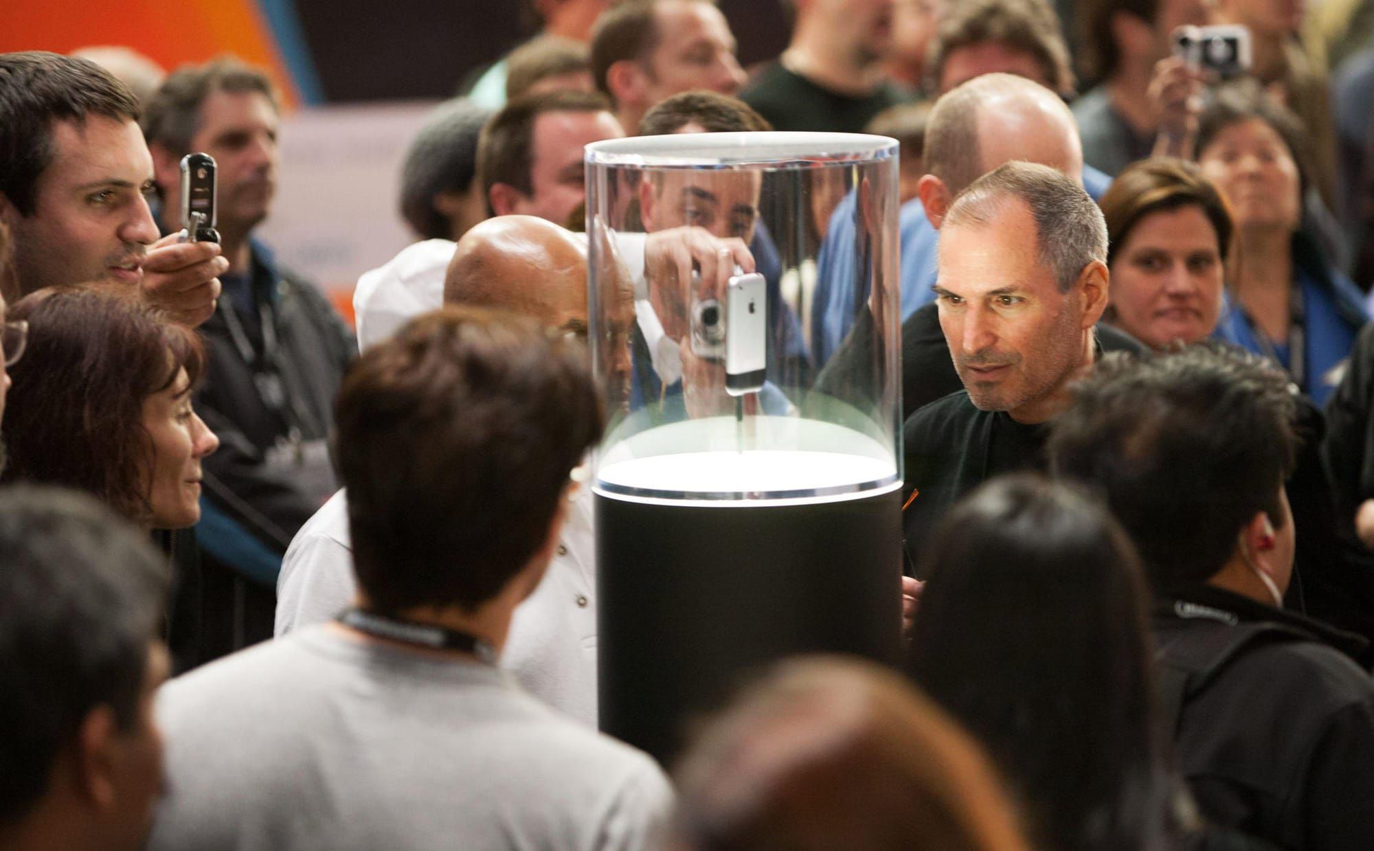 A big room with an enclosed first iPhone in the center, illuminated from the bottom. Steve Jobs is to it's right, looking at the phone, and a crowd surrounds both the podium and him.