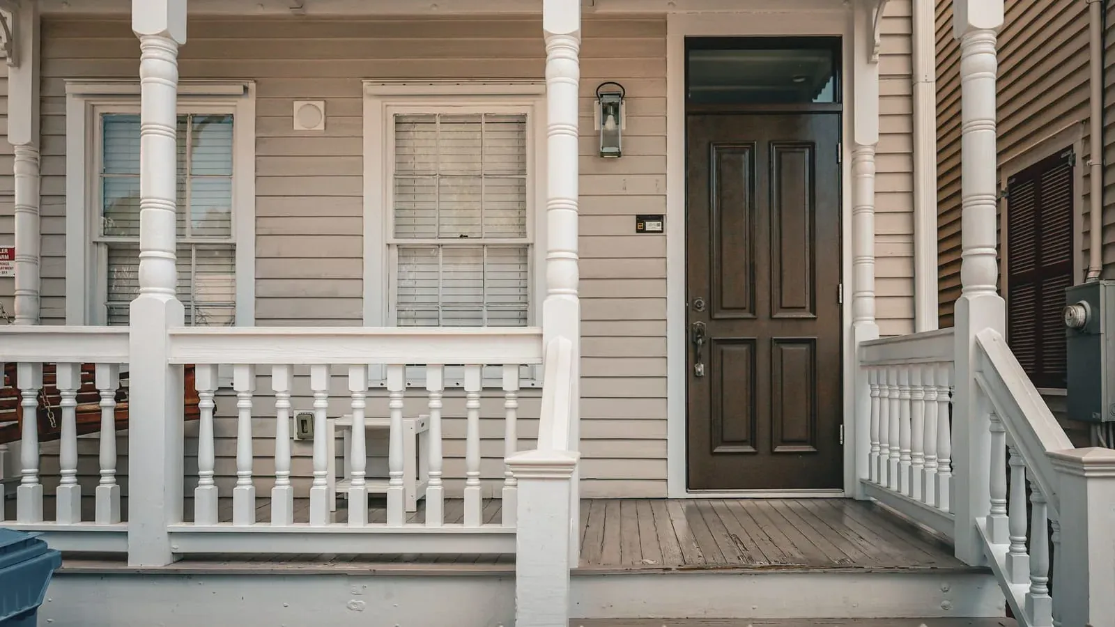 The front porch of a clapboard house, with turned white columns, a railing and a dark panelled front door