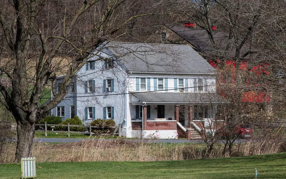 A large white farmhouse with blue shutters across a field, with paint damage visible near the porch