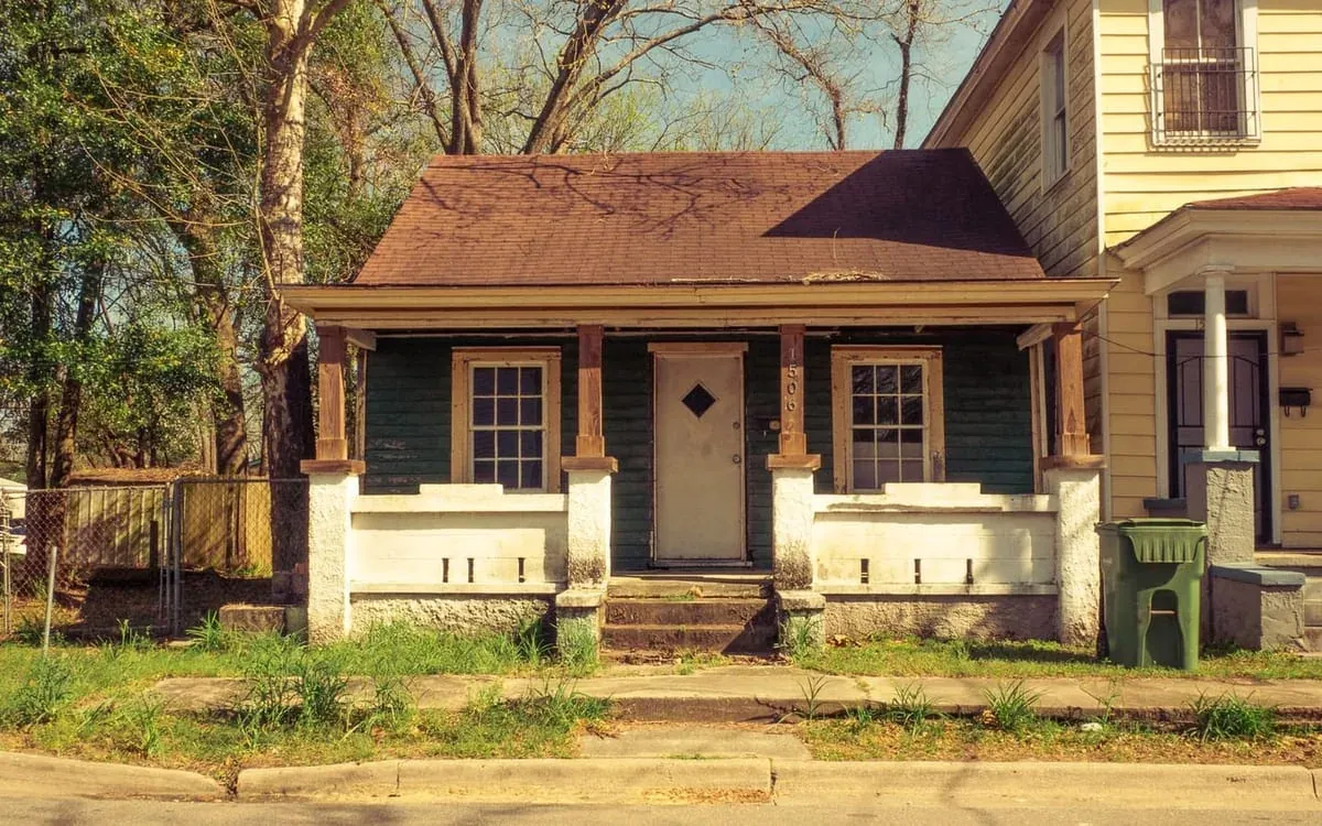 A small worn green cottage with a covered porch, an aged roof and weeds growing through the front path