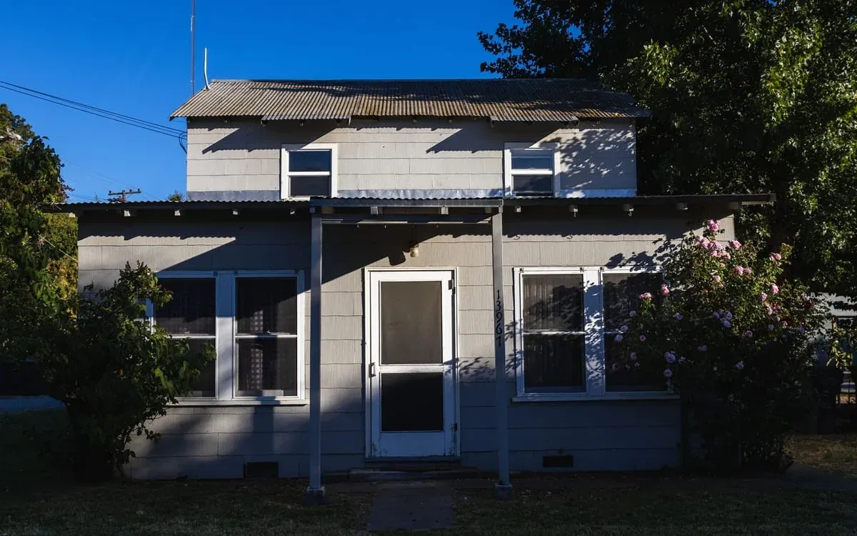 A small grey house with a corrugated metal roof and an aluminium porch awning, roses growing against one wall