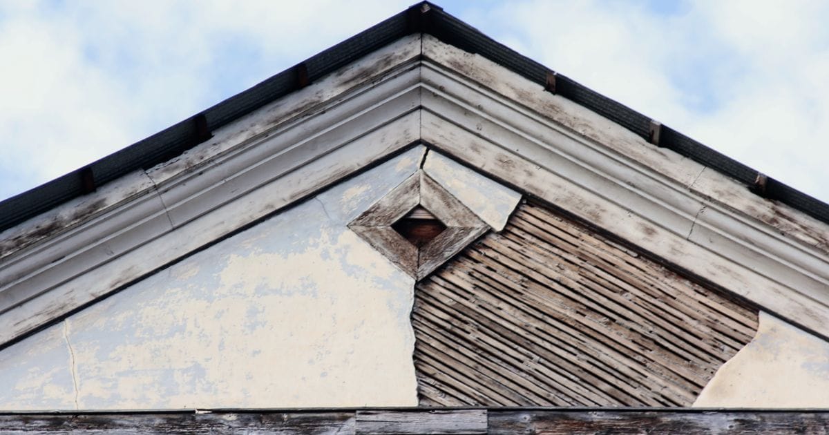 Gable end of an old building with cracked, peeling plaster exposing wooden lath beneath the roofline