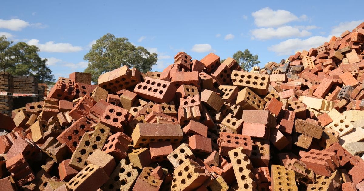 A large heap of broken red and tan bricks under a blue sky, with trees behind it