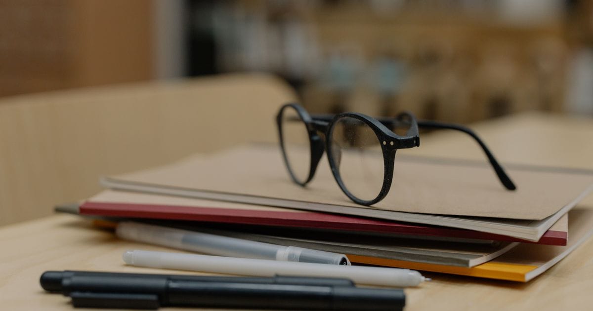 A pair of black reading glasses resting on a stack of folders and notebooks beside pens on a wooden table.