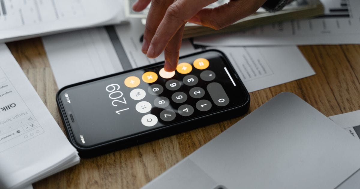 A hand tapping a calculator app on a phone lying on a wooden table covered with printed financial papers
