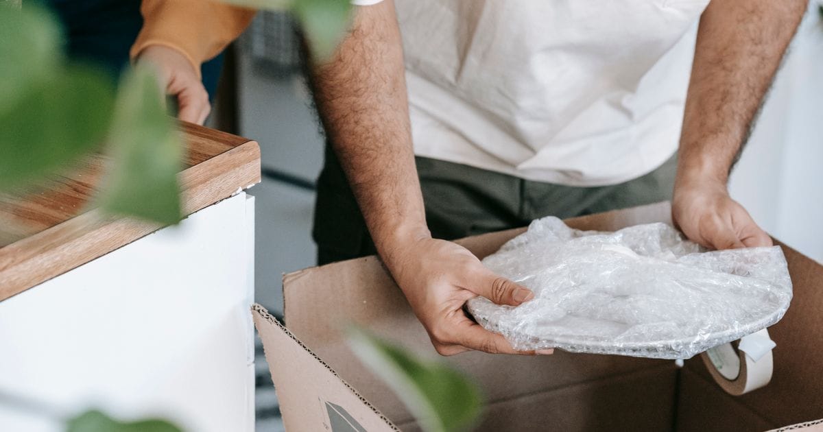 Hands placing a plate wrapped in bubble wrap into a cardboard box next to a kitchen counter