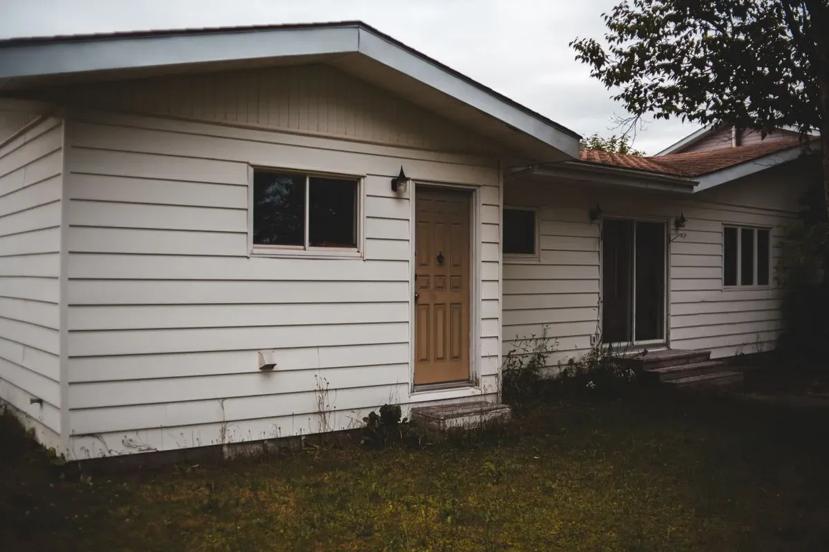 A plain single-storey house with white siding, a wooden front door and a patchy lawn