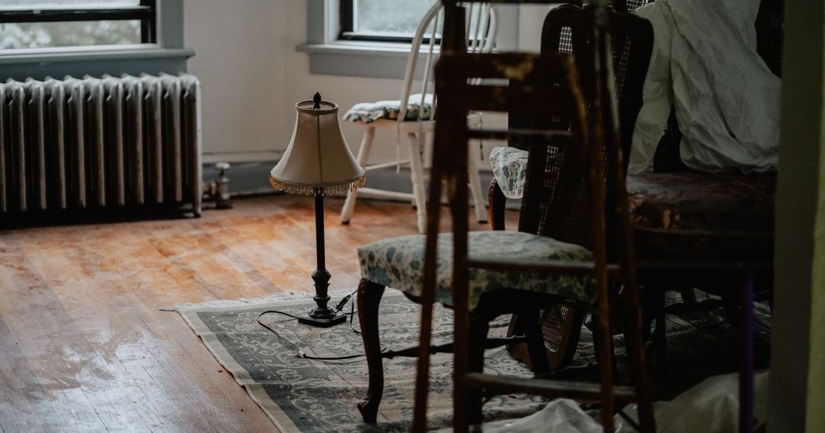 Room crowded with stacked wooden chairs and covered furniture beside a table lamp on a rug and a radiator under the window