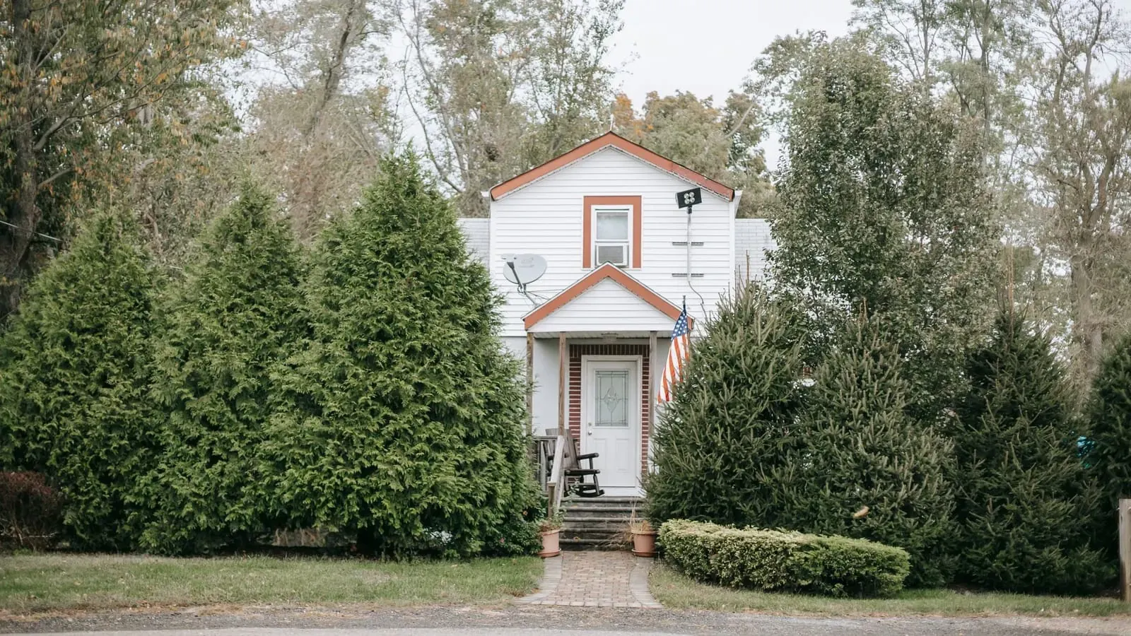 An older white clapboard house behind tall evergreens, with a small front porch and a satellite dish on the wall