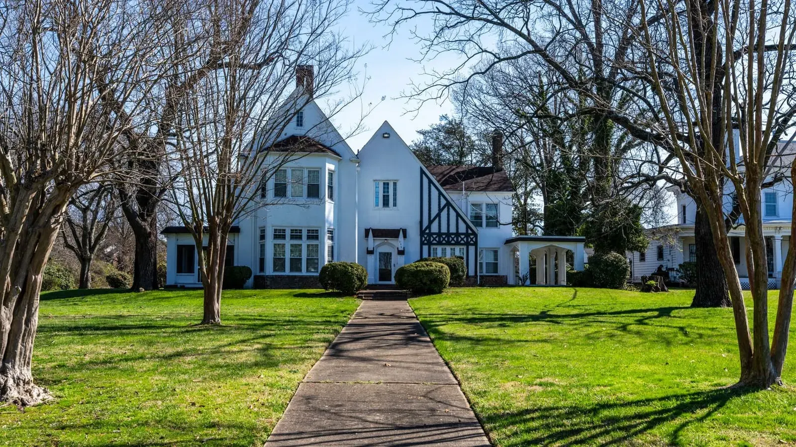 A white Tudor-style house with dark half-timbering and a side addition, on a large lawn with bare winter trees