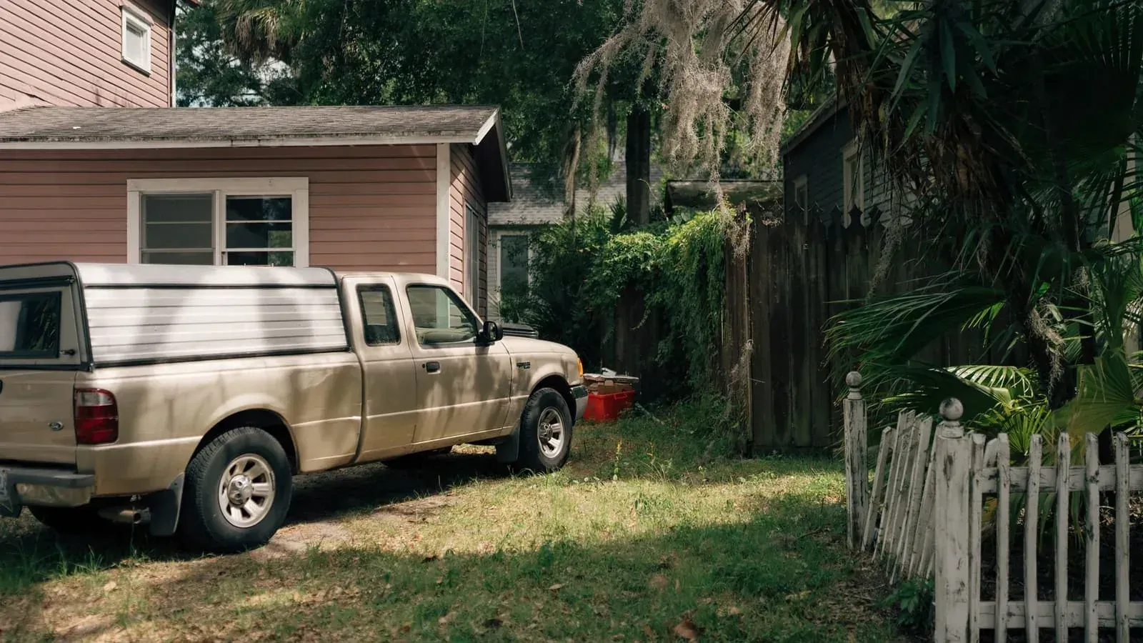 A pink clapboard house with a pickup truck parked on the grass, an overgrown yard and a leaning wooden fence