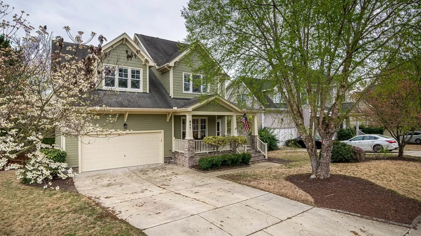 A green two-storey house with a two-car garage and a dogwood in flower, on a suburban street