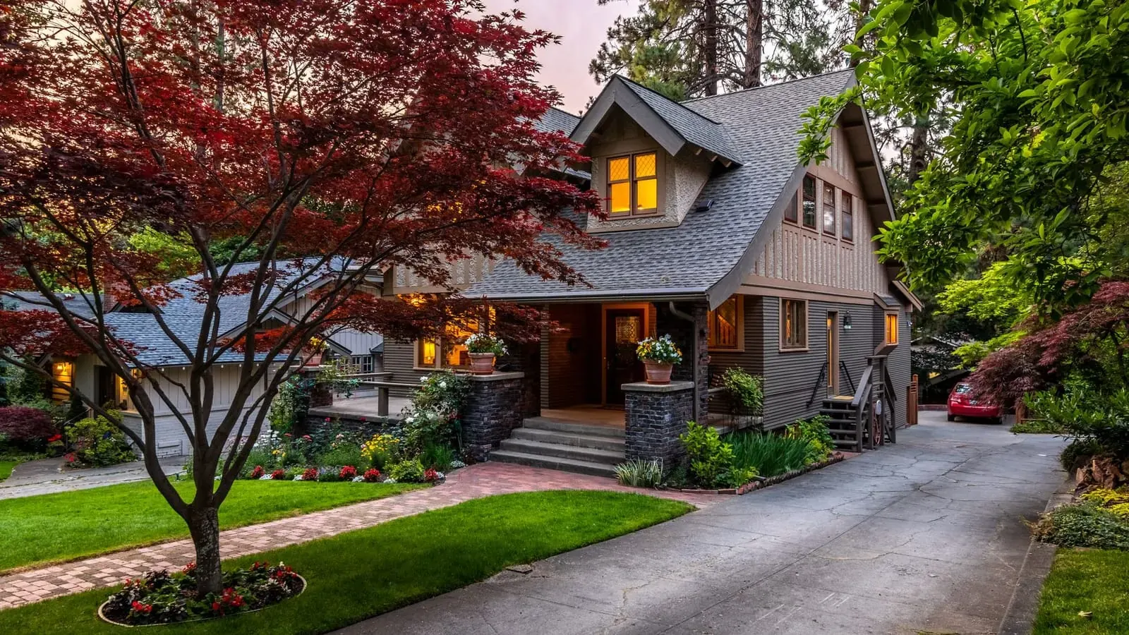 A craftsman-style house with a shingled roof and lit windows at dusk, a Japanese maple in the front garden