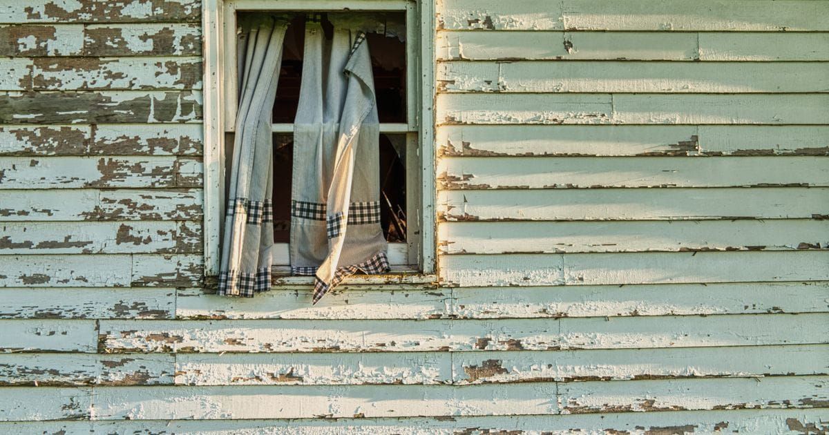 Pale green wood siding with flaking paint around a window with torn checked curtains