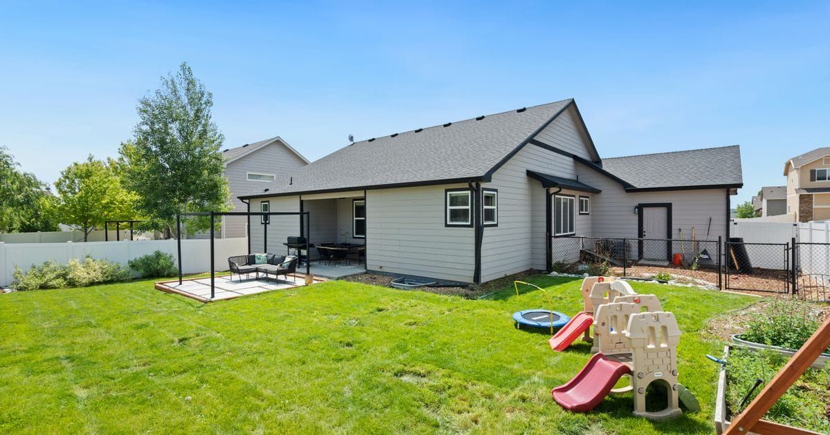 Back of a single-story gray house with a covered patio, a fenced green lawn and children's plastic slides in the yard