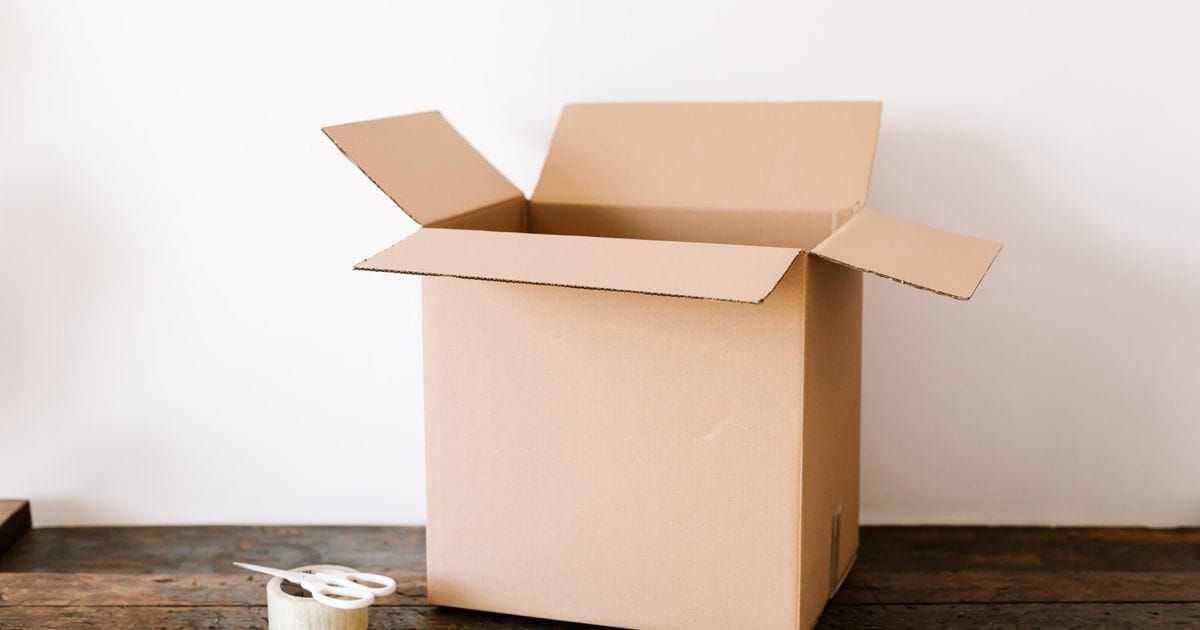An empty open cardboard moving box on a dark wooden table beside a roll of tape and white scissors