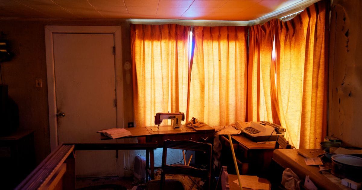 Cluttered room with a sewing machine, a typewriter and loose papers on wooden tables in front of orange curtains