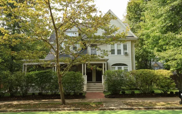 An older two-storey clapboard house with a covered front porch, set back behind a mature tree