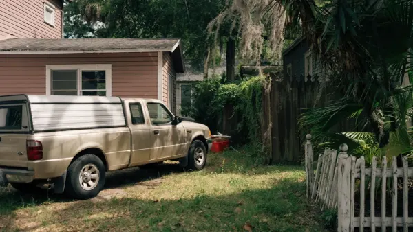 A pink clapboard house with a pickup truck parked on the grass, an overgrown yard and a leaning wooden fence