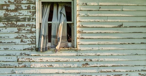 Pale green wood siding with flaking paint around a window with torn checked curtains