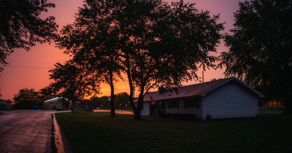 A single-story house with white siding beside large shade trees, a lawn and a wet street under an orange evening sky
