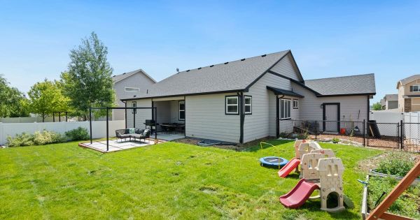 Back of a single-story gray house with a covered patio, a fenced green lawn and children's plastic slides in the yard