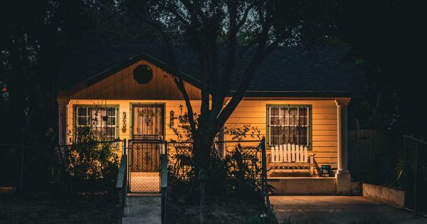 Small single-story yellow house with a front porch and a white bench, lit at dusk behind a tree and a low fence