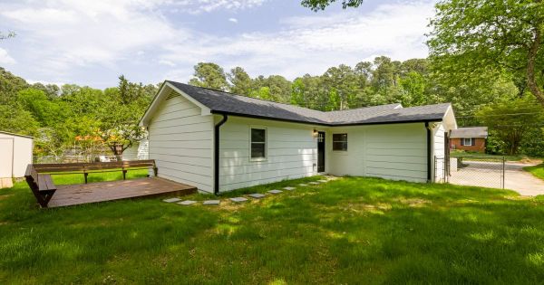 Single-story white house with dark shingle roof, a small wooden deck and a grassy yard under tall pine trees