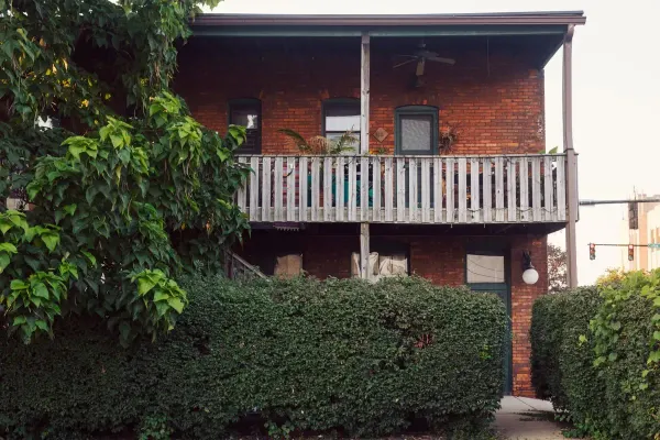 An older two-storey brick building with a weathered wooden balcony, partly hidden behind a tall overgrown hedge
