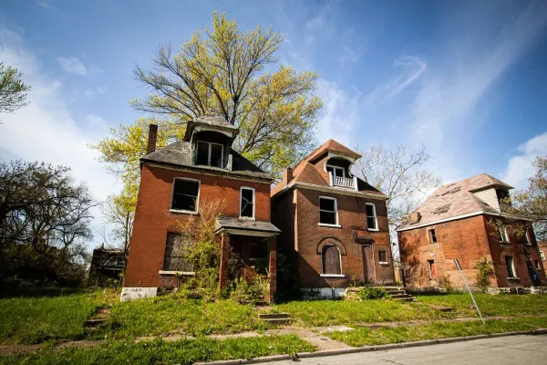Three empty brick houses with boarded and broken windows on an overgrown street, under a blue sky