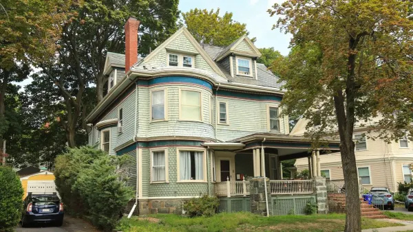 A tall older house with grey-green shingle siding, a red brick chimney and a wraparound porch, a car parked in the side driveway