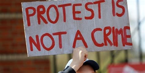 a protestor holds up a sign saying 'protest is not a crime' in red letters.