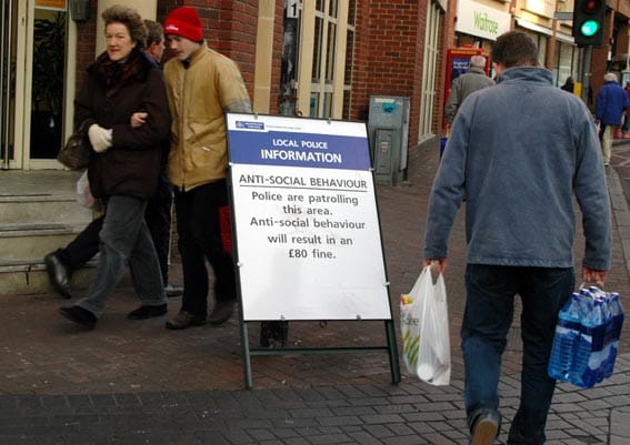 People walk past a police anti-social behaviour notice reading local poliice information: ANTI-SOCIAL BEHAVIOUR. Police are patrollomg this area, anti-social behaviour will result in an £80 fine. 