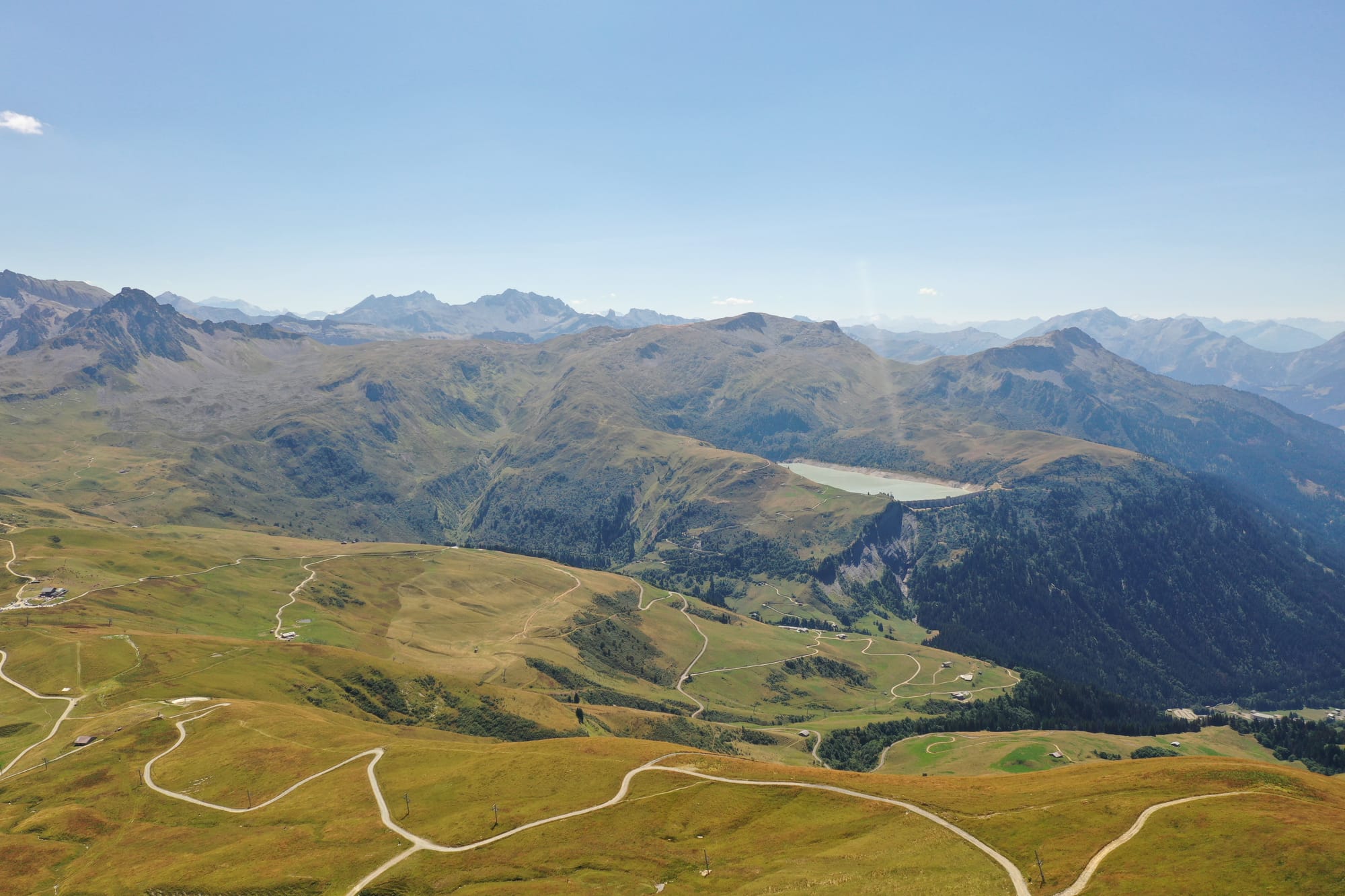 Aerial view of alpine pastures with winding trails and a turquoise mountain lake in the distance, captured with a DJI Mavic Pro 2 — above Megève, France