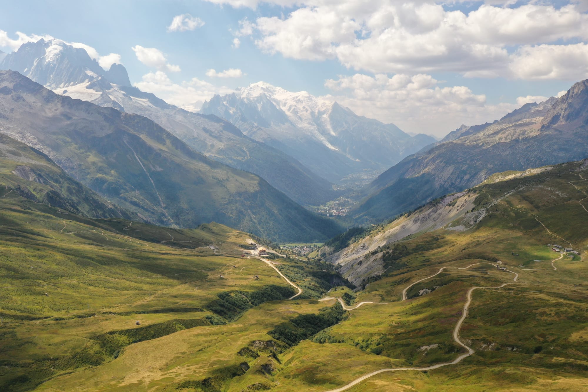 Aerial view of a vast green alpine valley with winding trails descending toward villages and the Mont-Blanc massif gleaming in the background, captured with a DJI Mavic Pro 2 — French Alps, France