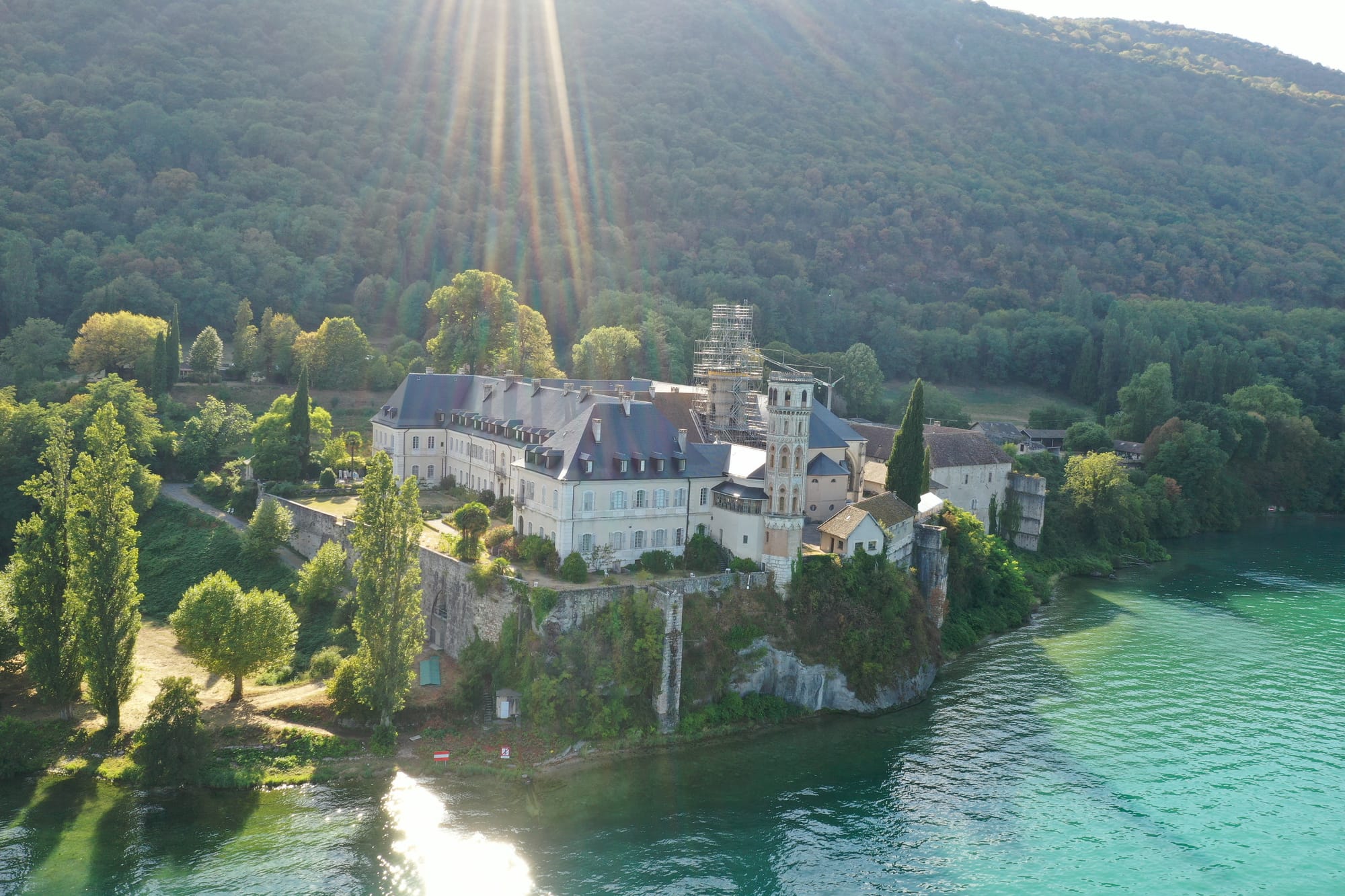 Aerial view of the Abbaye de Hautecombe on its lakeside promontory, surrounded by turquoise waters with golden sunlight streaming through clouds, captured with a DJI Mavic Pro 2 — Lac du Bourget, France