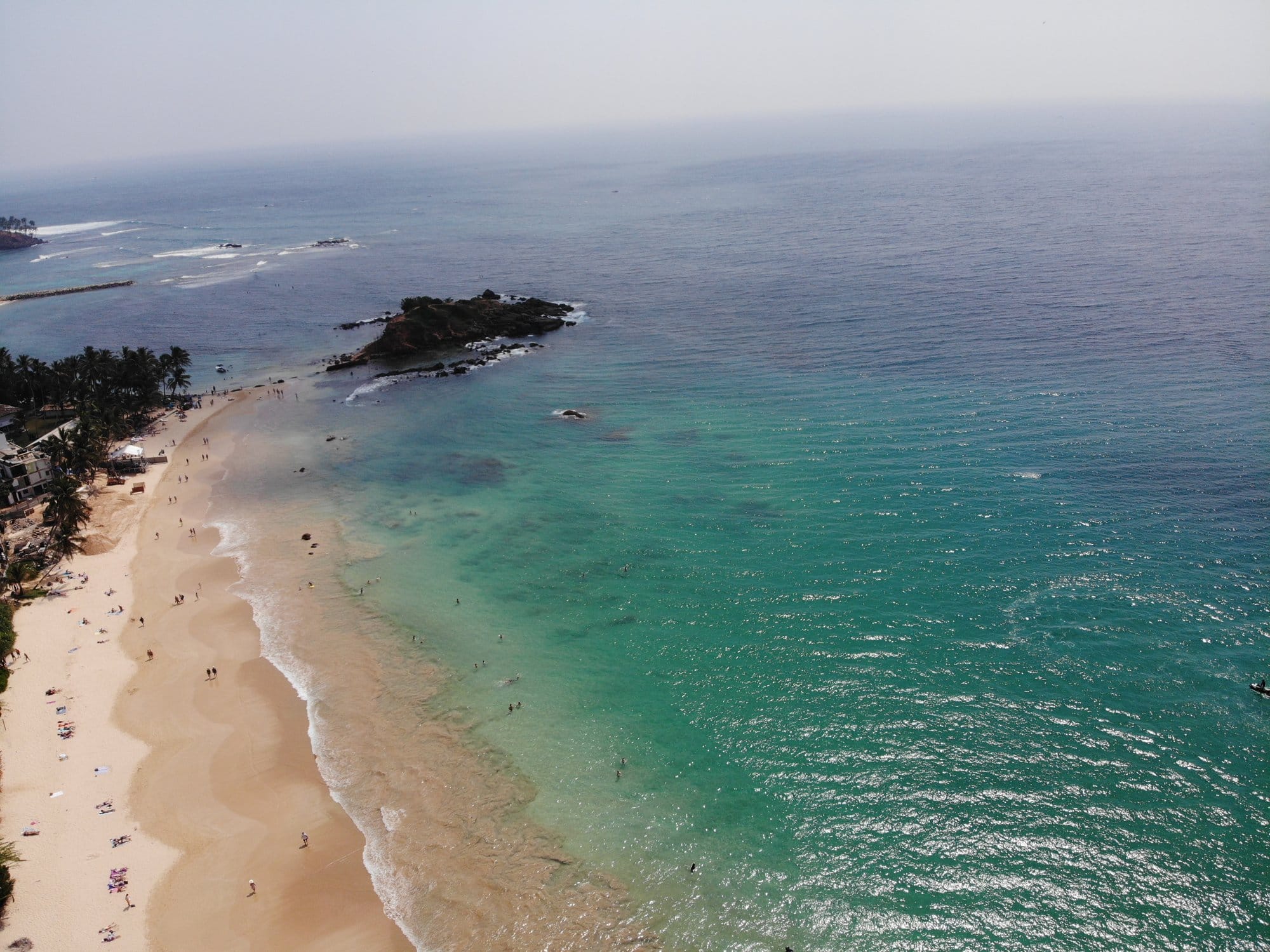 Crescent beach with crystalline turquoise water, swimmers and rocky island offshore, captured with a DJI Mavic Pro 2 — Mirissa, Sri Lanka