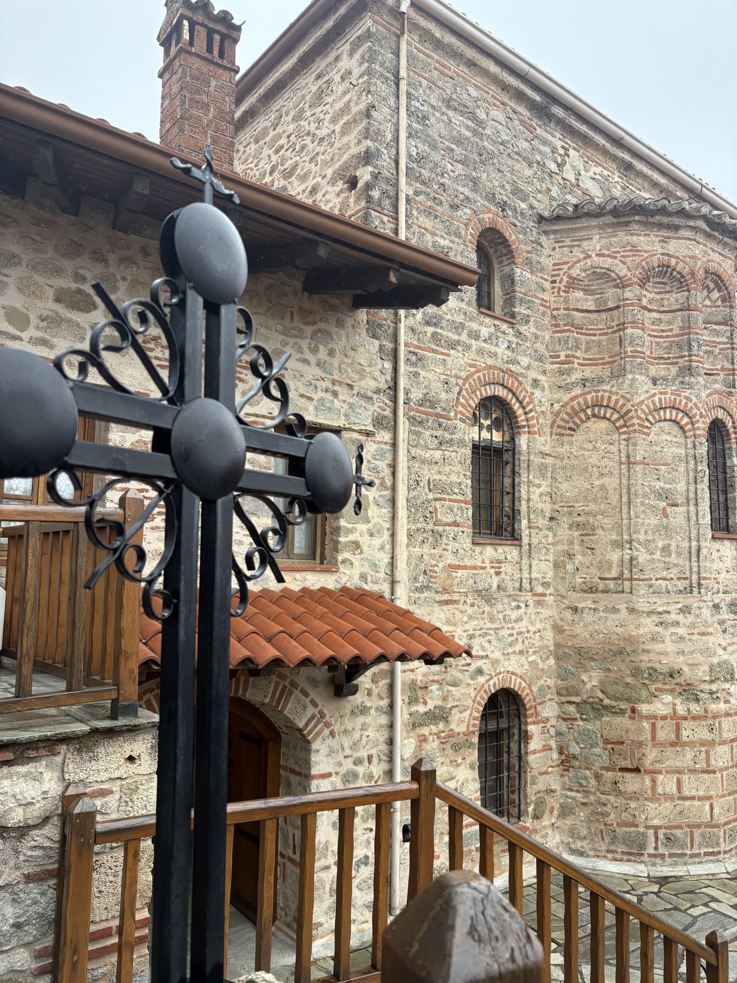 Ornate iron cross framing the stone-and-brick side wall of a Meteora monastery chapel — Greece