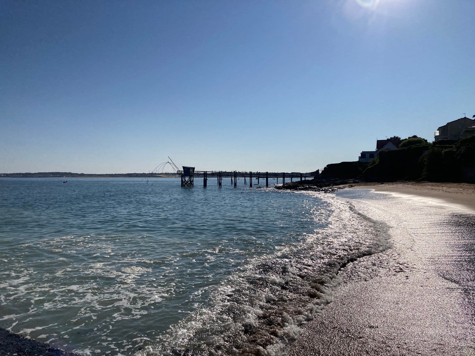Calm beach with gentle waves and carrelet fishing pier visible across the bay