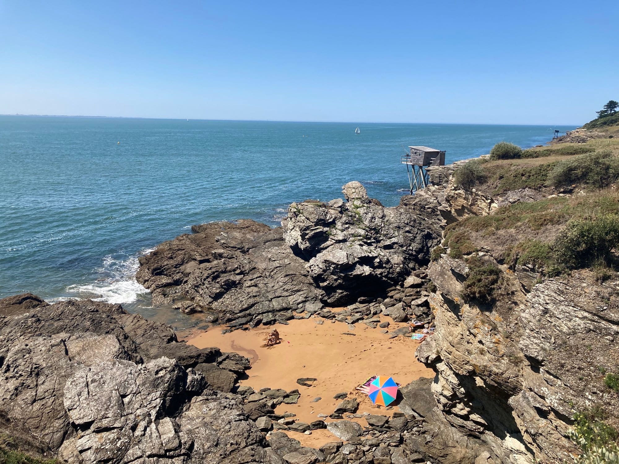Rocky coastal outcrop with a small sandy pocket beach and traditional carrelet hut on a pier in the background
