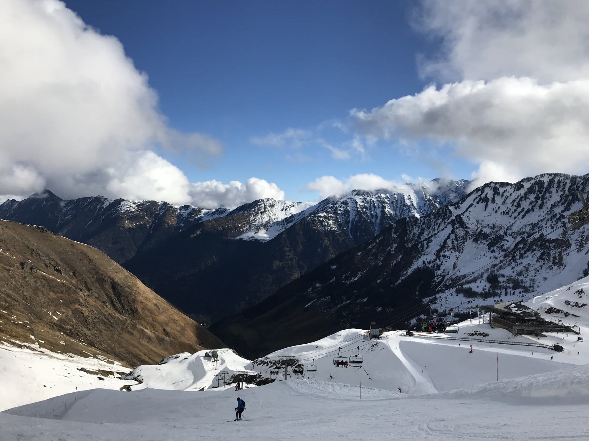A wide ski piste at the Cirque du Lys with a view down the valley — green slopes on the left, snow-covered peaks with clouds, and the resort buildings below — Cauterets, France