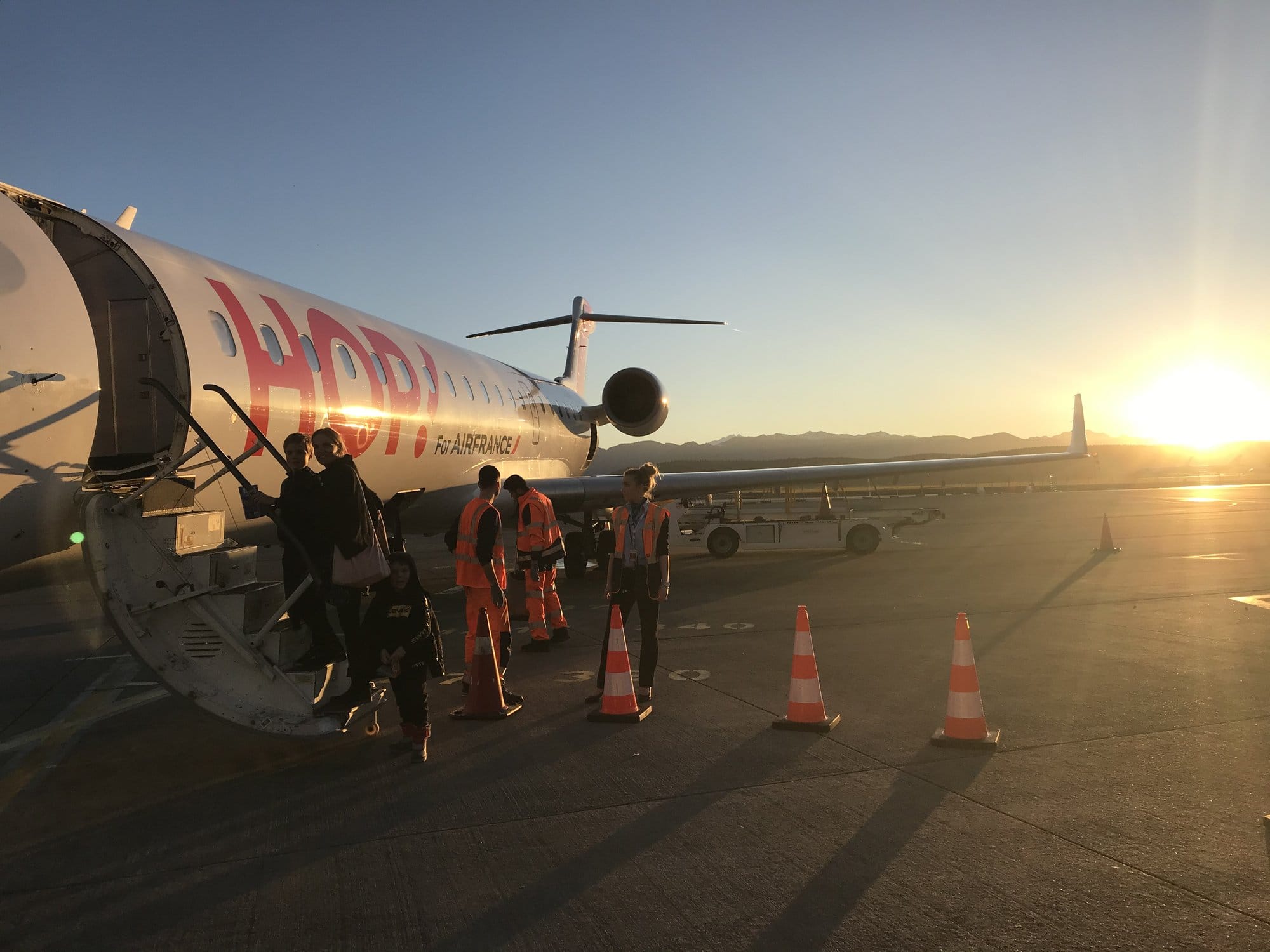 Passengers boarding a HOP! Air France plane on the tarmac at Tarbes-Lourdes airport at sunset, with golden light and orange cones — gateway to the Pyrenean ski resorts