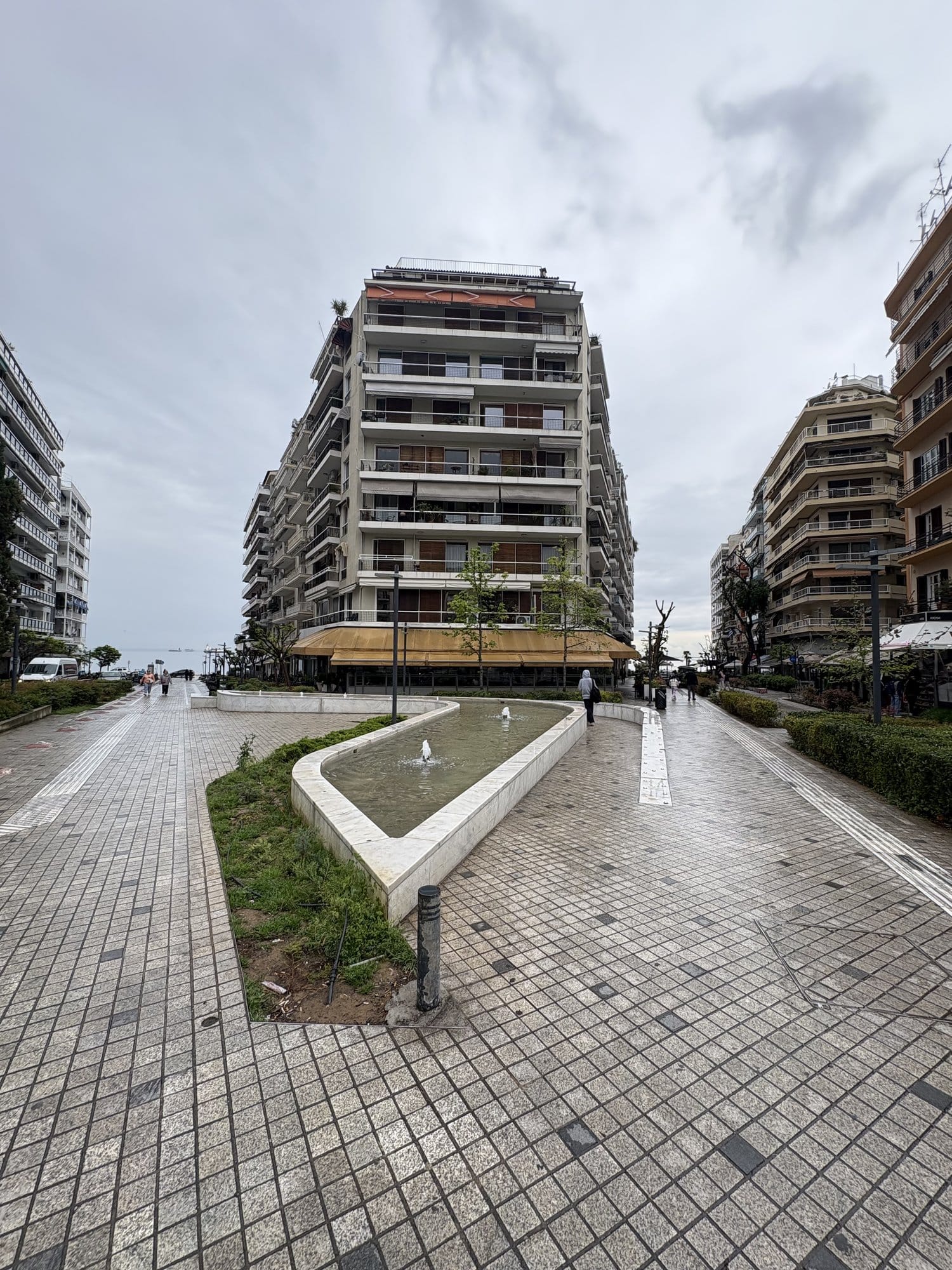 Promenade piétonne de Thessalonique avec un plan d'eau et un immeuble moderne, sous un ciel d'avril gris — Grèce