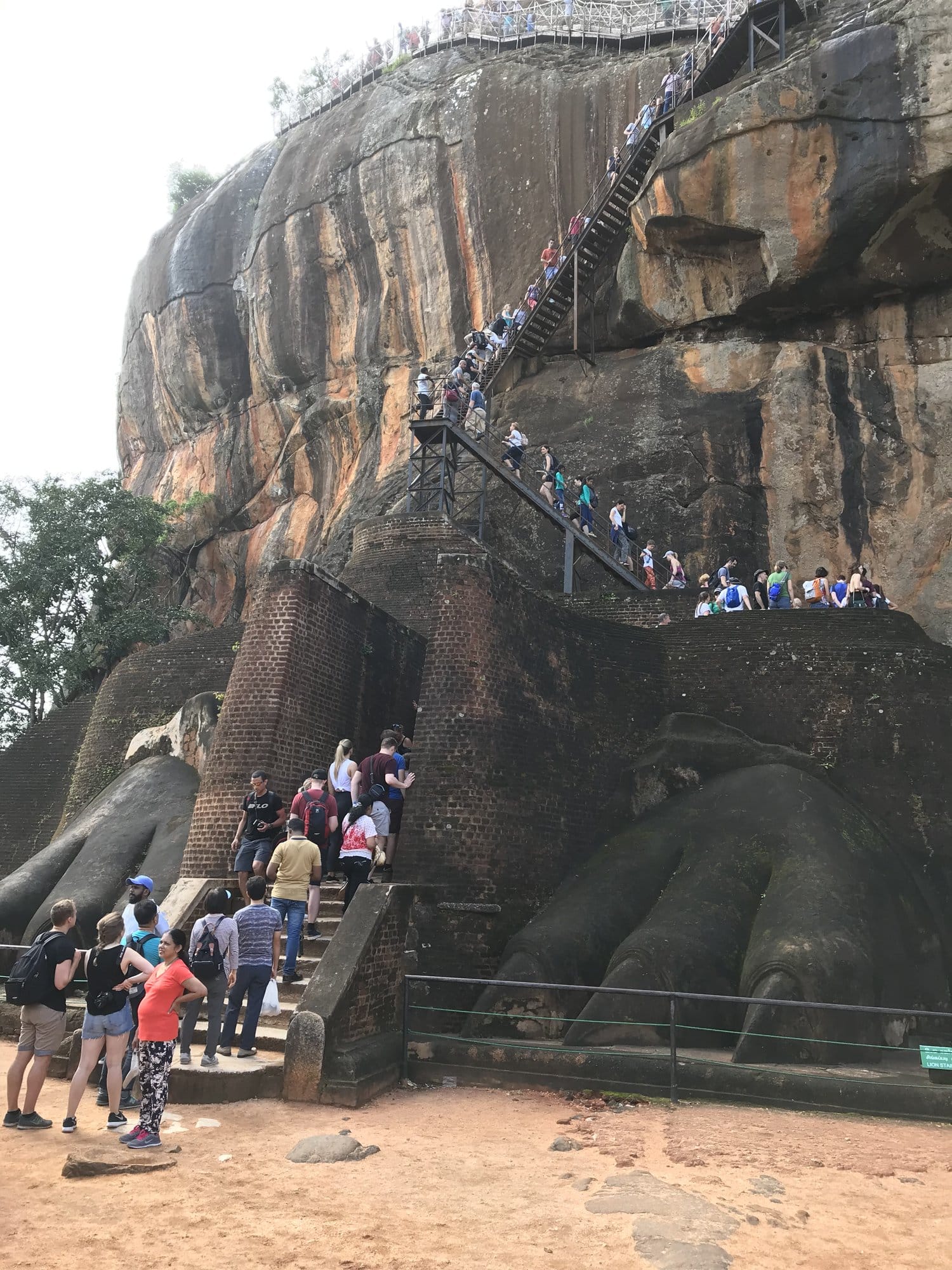 The metal staircase winds up Sigiriya's imposing rock face, visitors scattered across the weathered stone — Sigiriya, Sri Lanka