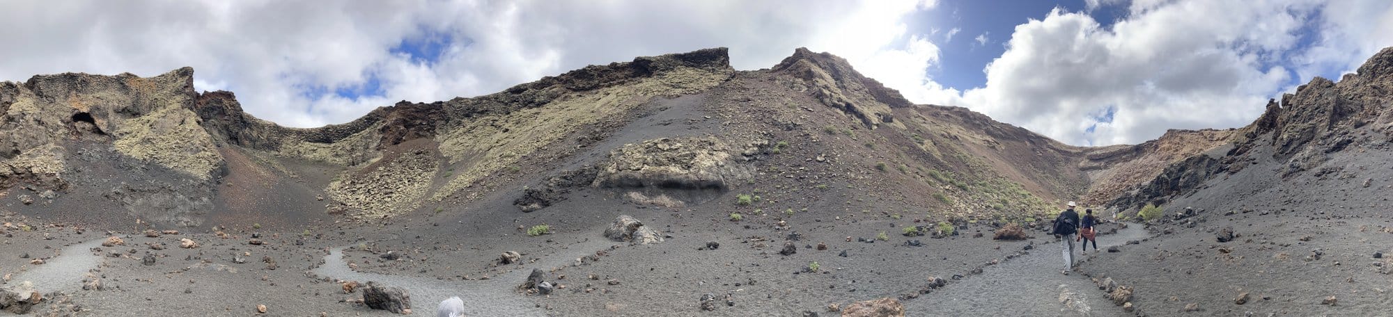 Panoramic view inside a volcanic caldera with hikers on the trail — the vast crater stretching across the frame with dark volcanic rock — Lanzarote