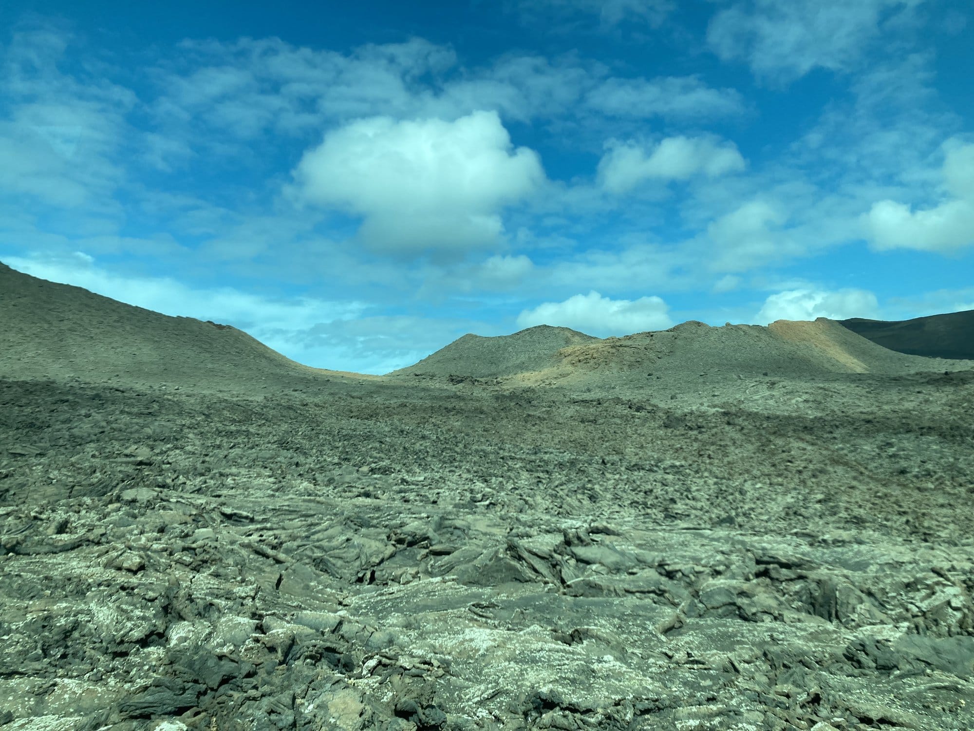 A vast field of solidified black lava stretching towards volcanic cones under a blue sky with scattered clouds — Timanfaya, Lanzarote